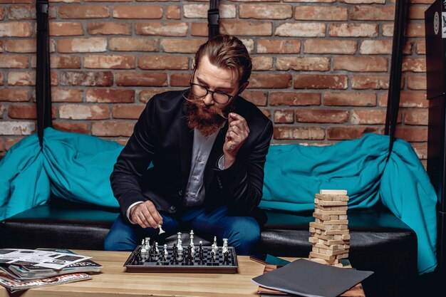 Intelligent handsome man with glasses, a beard and mustache, sitting on the couch and playing chess.