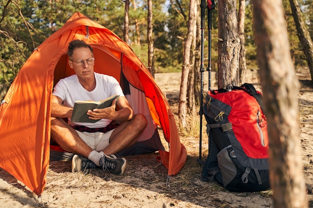 Intelligent mature male is sitting in tent and reading literature after nordic walking in forest