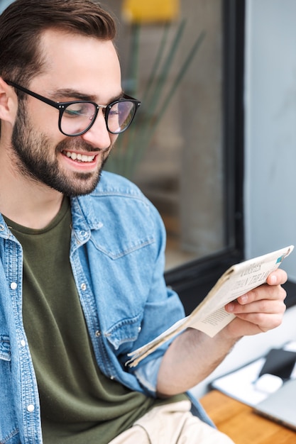 intelligente jonge man met een bril die lacht en de krant leest terwijl hij buiten in het stadscafé zit