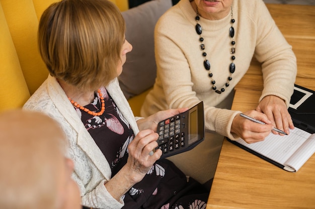 Intelligente knappe oudere vrouwen gekleed in beige en wit zitten in een modern café met oranje muren, rekenmachine en notitieboekje op tafel, discussiëren, tellen en cijfers opschrijven