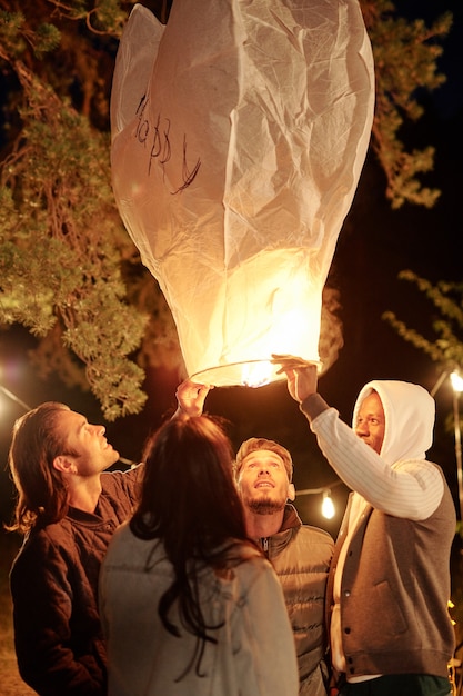 Interculturele jonge vrienden in vrijetijdskleding houden en kijken naar grote verlichte ballon 's nachts verzamelen onder dennenboom