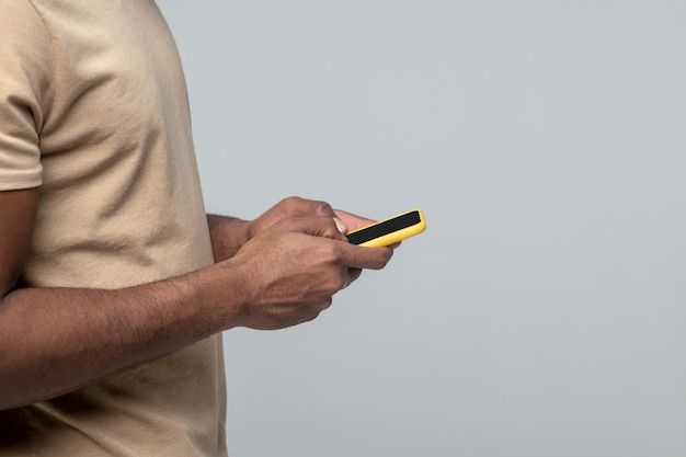 Interessant hier. Handen van Afro-Amerikaanse man in lichte tshirt permanent zijwaarts met smartphone op grijze achtergrond, zonder gezicht