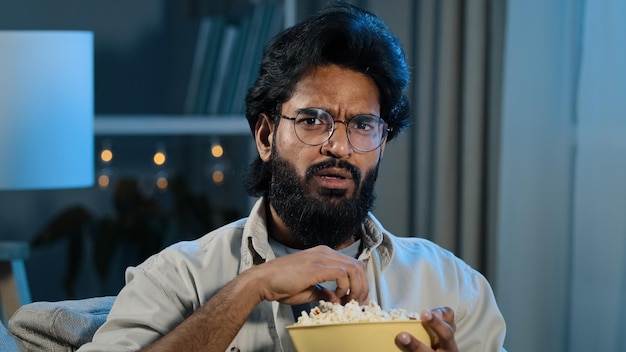 Interested attentive Arabic Hispanic Indian bearded man guy in glasses eating popcorn watching TV