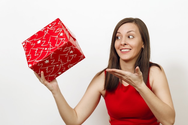 Interested and engaged young woman dressed in red dress and Christmas hat holds gift box, thinks that she was given on white background. Santa girl with present isolated. New Year holiday 2018 concept