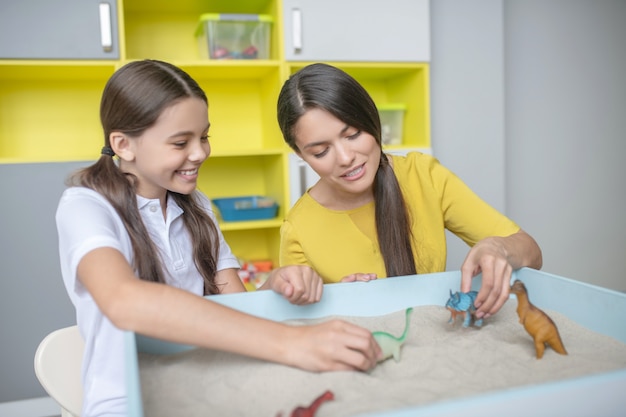 Interested joyful school-age girl and young woman playing with small animal figurines in sand tray