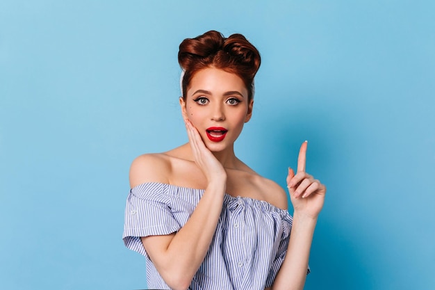 Interested pinup lady pointing up with finger Studio shot of sensual amazed girl isolated on blue background