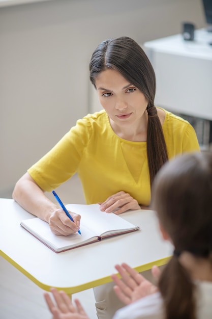 Interested smart beautiful woman in yellow blouse sitting at table with pen and girl in front