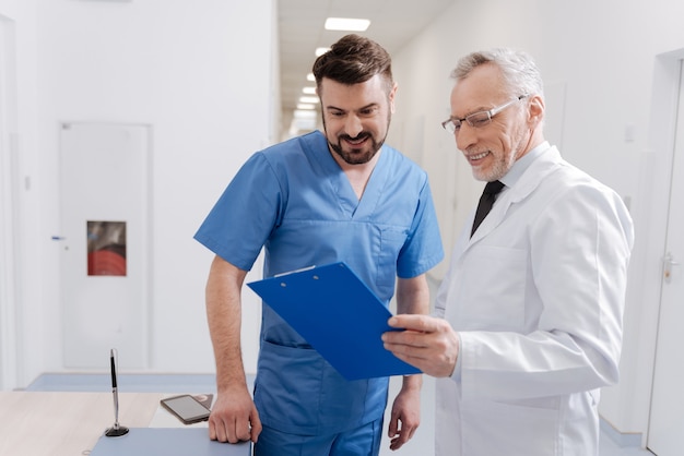 Interested in your opinion. Optimistic skillful old pediatrician working in the hospital and signing statement while senior colleague talking on the phone in the background