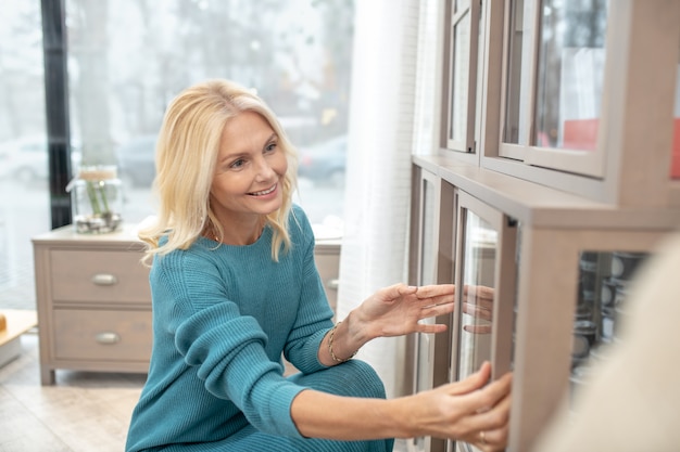 Interesting design, cupboard. Interested woman crouched near a glazed cupboard, opening a small door with her hands, in delight.