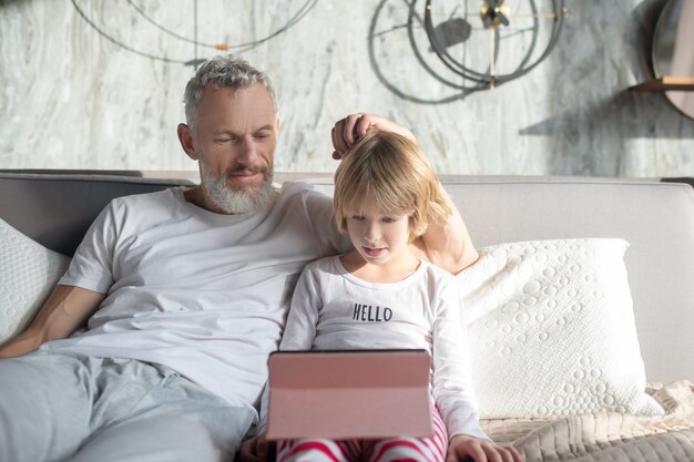 Interessante qui. attento papà interessato e bambino guardando lo schermo del tablet seduto sul letto di casa