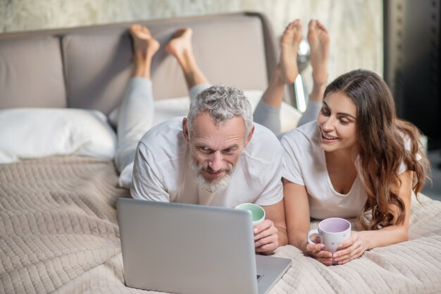 Interesting here. Smiling concerned bearded man and long-haired woman with coffee lying in front of laptop on bed