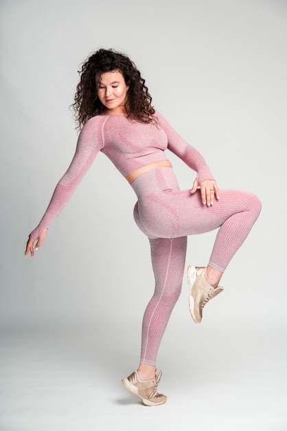 Interesting pose. Full length view of the brunette curly woman standing at one leg and looking down while posing over the white wall background