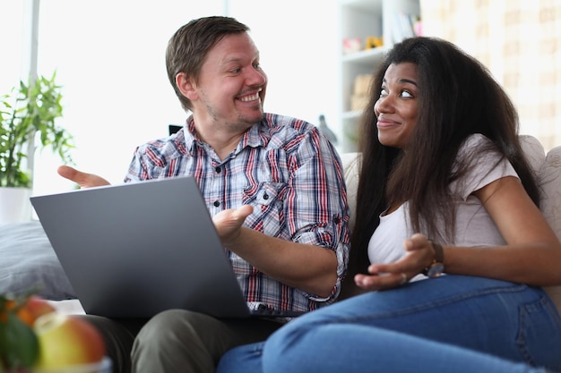 Interethnic family couple laughing and chatting while sitting on sofa at home with laptop