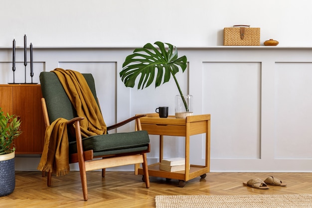 Interieur van stijlvolle woonkamer met vintage groene fauteuil, houten salontafel, meubels, grijze muur, plank, tapijt, planten, bladeren in vaas, boek, kopieerruimte en elegante persoonlijke accessoires.