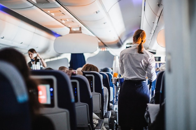 Интерьер самолета с пассажирами на сиденьях и стюардессой в форме, идущей по проходу, обслуживающей пе