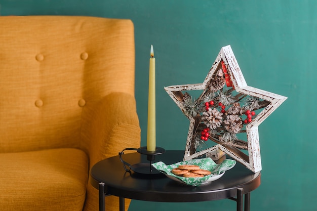 Photo interior christmas photo with burning candles. next to a vintage yellow sofa against a dark green wall on the table there is a christmas star, a burning candle and a plate of cookies.