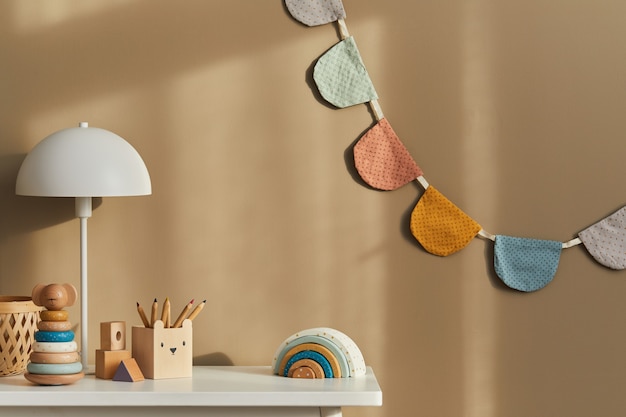 Design degli interni di un'elegante stanza per bambini con mensola bianca, giocattoli in legno, accessori per bambini, lampada bianca, decorazioni accoglienti e bandiere di cotone appese al muro beige.