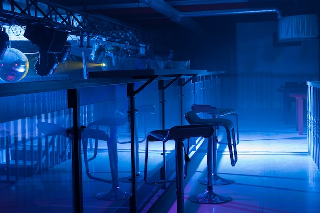 Interior of a modern bar with moody blue lighting and a row of contemporary bar stools in front of a reflective counter in a nightclub