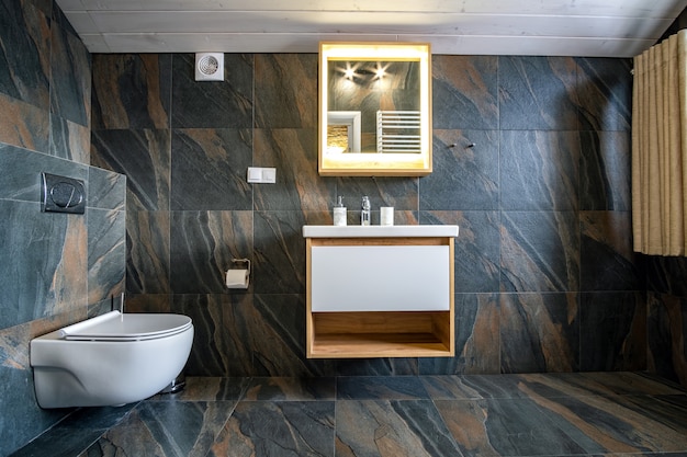 Interior of modern stylish bathroom with black tiled walls, curtain shower place and wooden furniture with wash basin and big illuminated mirror.