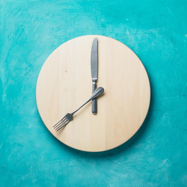 Intermittent fasting and skip breakfast concept  empty wooden round tray or trencher with cutlery as clock hands on blue background Eight hour feeding window concept or breakfast time concept