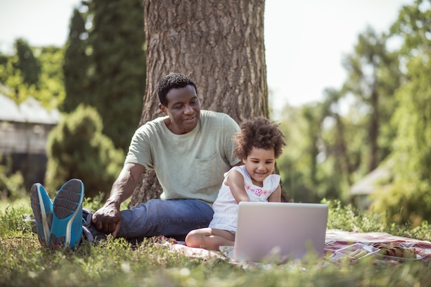 On internet. Curly-haired little girl sitting under the tree with her dad and watching something online