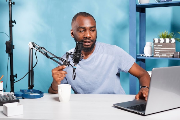 Internet influencer speaking in microphone and using laptop while recording video for blog in home studio. African american vlogger streaming live talking and reading from computer in mic