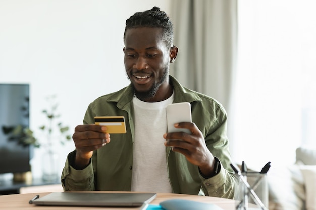 Internet mobile shopping Happy black man using cellphone and credit card purchasing online while sitting at desk