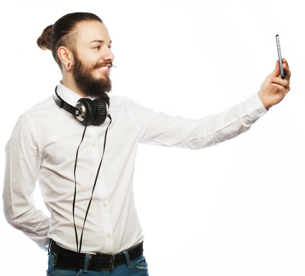 Internet tehnology and people concept a young man with a beard in shirt holding mobile phone and making photo of himself while standing against white background