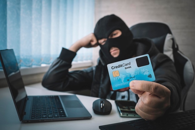 사진 인터넷 절도 한 남자가 발라클라바를 착용하고 노트북 흰색 배경 뒤에 앉아 있는 동안 신용 카드를 들고 있습니다.