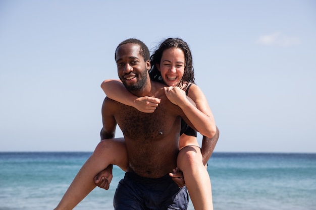 해변에서 인종 간 부부, 아프리카 남자가 백인 여자를 들어 올려 여름에 그들은 휴가를 즐기고 재미를 느낍니다.