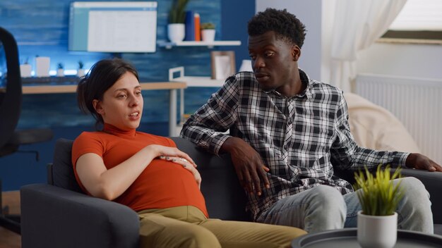 Interracial couple expecting child in living room while talking about baby and parenthood. Multi ethnic people with pregnancy sitting together on couch at home and bonding over child