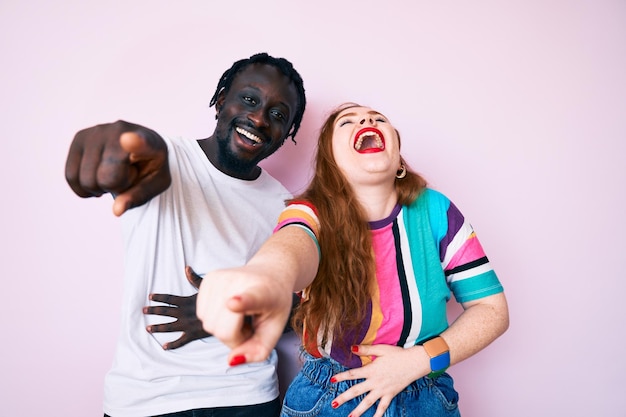 Interracial couple wearing casual clothes laughing at you pointing finger to the camera with hand over body shame expression