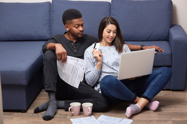 Interracial couple of young people sitting together in front laptop on floor near sofa, in their appartments