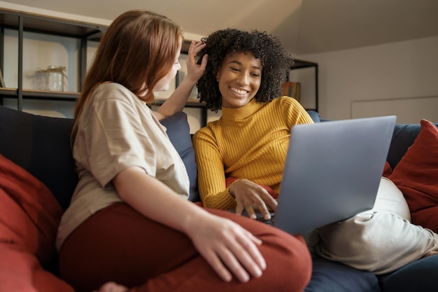 Coppia di donne gay interrazziali che usano il laptop e si divertono seduti sul divano