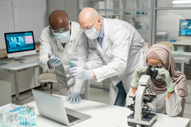 Interracial male scientists discussing blue liquid in flask next to colleague