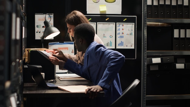 interracial police partners conduct detective work, examining evidence in archive room, searching for clues to solve a crime. serious female inspector assisting focused male detective with case files.