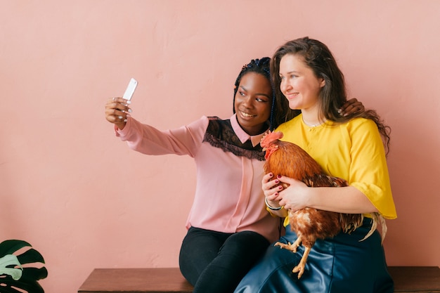 Interracial women with chicken in hands make selfie over pink wall