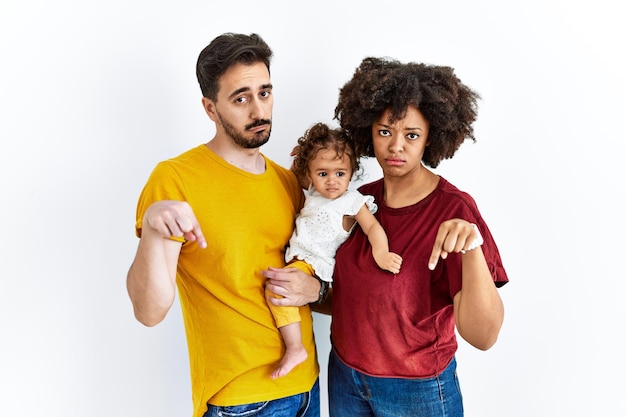 Interracial young family of black mother and hispanic father with daughter pointing down looking sad and upset indicating direction with fingers unhappy and depressed