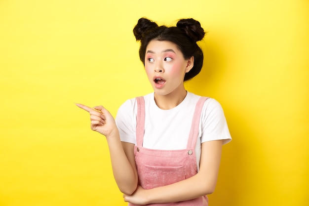 Intrigued asian beauty girl with makeup, looking curious at logo, pointing finger aside, standing in summer clothes against yellow.