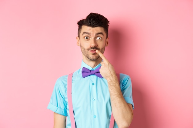 Intrigued happy guy touching lip, looking at camera pensive, making choice what to buy, standing over pink.