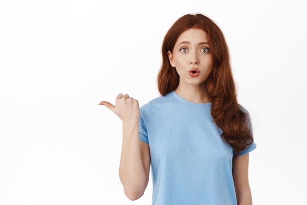 Intrigued redhead natural girl say wow, pointing finger left and look at camera with surprised and interested face, ask question about special promo deal in shop, white background