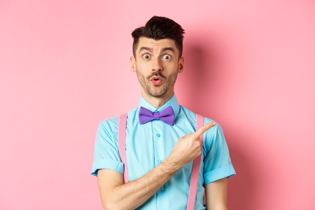 콧수염이 멋진 프로모션을 보여주고 호기심을 보이고 와우라고 말하고 로고에서 손가락을 가리키며 분홍색 배경에 서있는 흥미롭고 놀란 귀여운 남자.