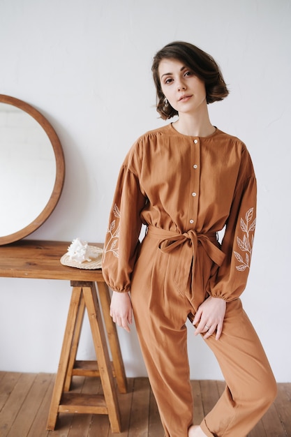 Intriguing woman possing for a photo in a brown jumpsuit, looking at the camera. Beautiful brunette with short hair indoors, next to a table with a round mirror