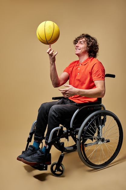 Invalid man spin the ball on finger, smiling, having fun alone while sitting on wheelchair, isolated over beige background