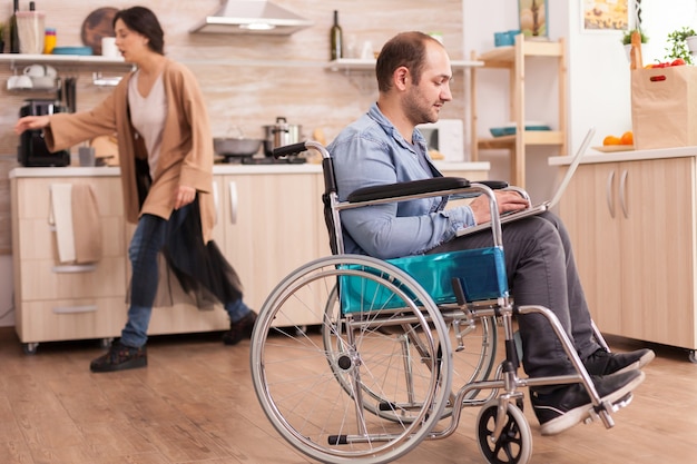 Invalid man in wheelchair working remotely on laptop in kitchen while is standing in front of refrigerator. Disabled paralyzed handicapped man with walking disability integrating after an accident.