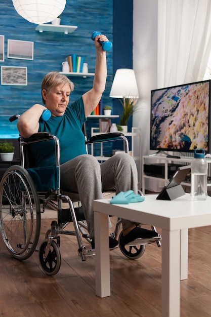 Invalid senior woman holding workout dumbbells raising arm while exercising body muscle persistence ...