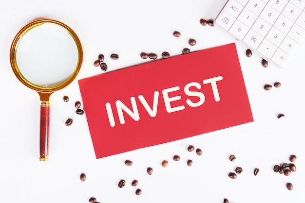 Invest word on a red card with scattered coffee beans on the table and a magnifying glass as a symbol reminding you to focus when choosing an investment