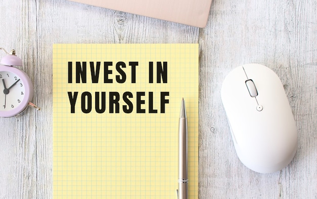 INVEST IN YOURSELF text written in a notebook lying on a wooden work table next to a laptop