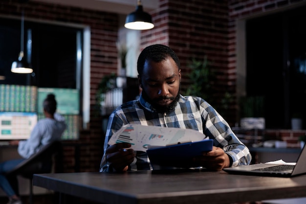 Investment company analyst reviewing market trend documentation while sitting at desk in office at night. Forex stock agent analyzing financial charts while looking at paperwork clipboard.