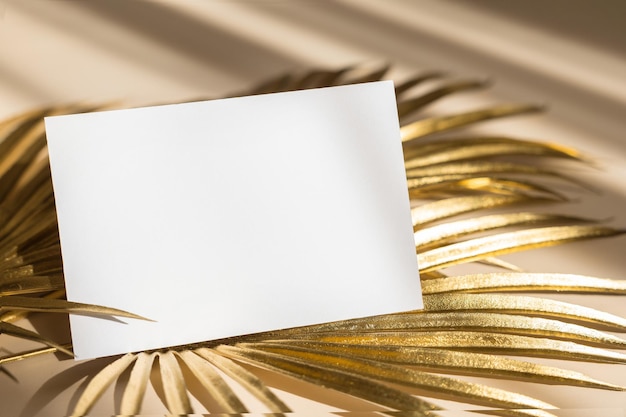 ベージュのパステルカラーの背景に金色のヤシの葉の招待状のモックアップ上面図フラットレイコピースペースブランディングと広告のための白い紙のモックアップのテンプレート空白